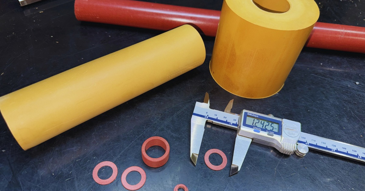 Several Rulon® rings sit on a black surface, next to a measuring tool. Yellow and red cylinders sit nearby. | Plastic Machining Inc. Several Rulon® rings sit on a black surface, next to a measuring tool. Yellow and red cylinders sit nearby.