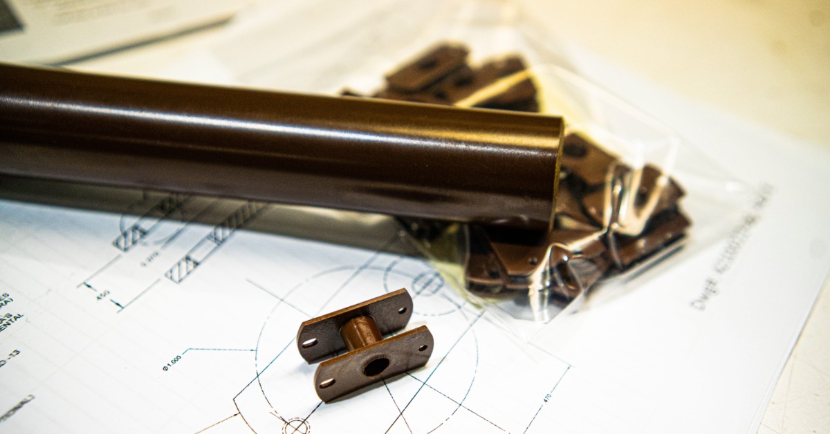 A long brown Vespel® tube sits on a piece of paper showing plans, along with a bag of small components.