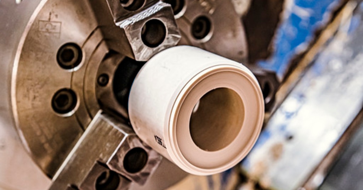 PEEK material secured in a metal lathe chuck, with blue machinery and blurred workshop surfaces nearby.