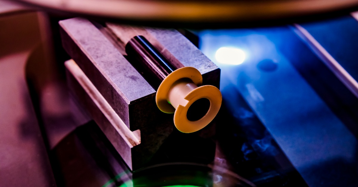 A yellow and black cylindrical part clamped in a metal fixture beside a blue work surface and bright light. | Plastic Machining Inc. A yellow and black cylindrical part clamped in a metal fixture beside a blue work surface and bright light.