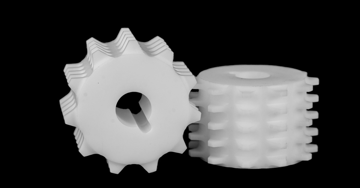 Two white plastic gear-shaped parts with ridged edges and center holes displayed on a black background. | Plastic Machining Inc. Two white plastic gear-shaped parts with ridged edges and center holes displayed on a black background.