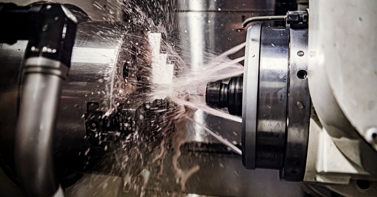 A metal machining tool cutting a part while liquid sprays across silver machinery inside an industrial machine.