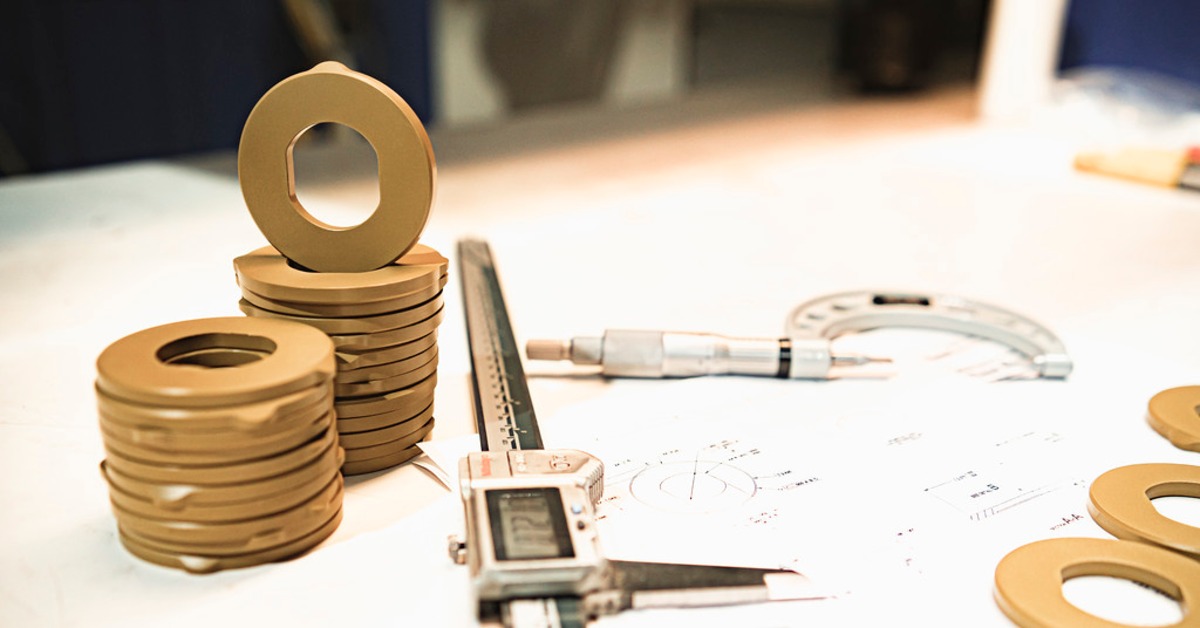 7 Best Plastics for Continuous High Heat Exposure | Plastic Machining Inc. Stacks of tan ring-shaped parts beside digital calipers, a micrometer, and technical drawings on a worktable.